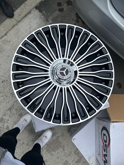 An overhead, medium shot captures a person standing next to a Mercedes-Benz wheel. The wheel is the primary focus and is positioned in the center of the frame. It features a chrome Mercedes-Benz logo in the center, surrounded by a glossy black rim with thin chrome accents. The wheel is designed with numerous spokes that create a dynamic, multi-dimensional pattern.
The person is wearing black pants and white sneakers. Their feet are visible at the bottom of the frame, indicating they are standing on a concrete surface. Adjacent to the wheel, there is a cardboard box with "DSW WHEELS" branding. In the upper left corner, a dark shoe is visible on the concrete. The background includes a portion of a silver car and the concrete ground, suggesting an outdoor setting, possibly a driveway or sidewalk. The lighting suggests it is daytime. The scene does not provide any explicit clues about the location in Teteven, Bulgaria.