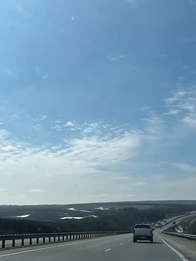 The image shows a view from a vehicle moving along a highway in the direction of Russkie Kirmeni, Russia. The sky is blue with wispy clouds. To the right of the highway is a forested area with patches of snow. The highway has multiple lanes, a metal safety barrier, and white lane markings. Another vehicle is visible to the right on the road.