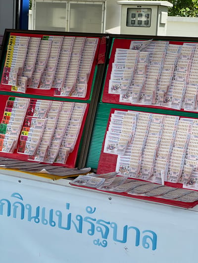 The image shows a lottery ticket stall in Tambon Phraek Ha, Thailand. The stall displays a large number of Thai lottery tickets, neatly arranged in several rows. The tickets have various numbers and designs, typical of lottery tickets. In the background, there is a white utility box with several dials and indicators, possibly an electrical or communication equipment box. Partially visible in the foreground is a blue sign with Thai text, suggesting it is part of the stall's branding or advertisement. The lighting suggests it is daytime, and the overall environment appears to be outdoors. There are no people visible in the image, and the focus is on the lottery tickets and the stall's setup.