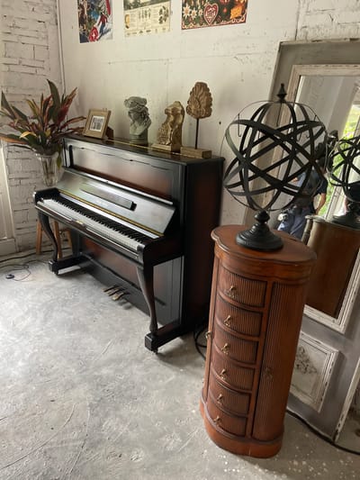 The image depicts an indoor scene with a dark wood upright piano as the central focus. The piano is positioned on a grey concrete floor with visible scuff marks and textures. To the left of the piano, a vase filled with a colorful, leafy plant stands near a white brick wall. On top of the piano, a small framed photograph and two decorative busts are visible. To the right of the piano, a tall, narrow, wooden cabinet with five drawers and small brass pulls is present. This cabinet supports a black metal armillary sphere. In the background, a large, ornate mirror leans against the wall, reflecting a person wearing a blue shirt and dark pants. The walls appear to be rough-textured and painted white, with some decorative posters or prints affixed near the top. The overall ambiance suggests a curated interior space, possibly a showroom, studio, or antique shop. No specific landmarks or street views are visible to indicate the location in Guangzhou City, China.
