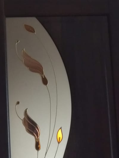 The image is a close-up of a decorative glass panel, possibly part of a door or window. The panel features an abstract floral or leaf design in muted colors, with accents of metallic brown and a small, bright yellow element that resembles a flame or a dewdrop. The design is set against a pale, off-white background. To the right of the glass panel is a dark, textured surface, likely wood, which forms the surrounding frame or structure. The lighting suggests an indoor environment, with the yellow element appearing illuminated from behind. The image does not contain any people or clear indicators of its specific location in Otaci, Moldova.