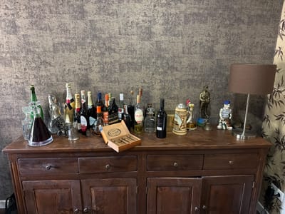 The image depicts a wooden sideboard against a textured, mottled brown and grey wall, likely in Bissendorf, Germany. On the top of the sideboard, a collection of assorted alcoholic beverages in various bottles and decanters are displayed. Notable among the bottles are several champagne bottles, a red wine bottle, and various spirits. To the right, a dark brown lampshade sits atop a silver lamp base. Several decorative figurines are also present, including a soldier, a man in a striped shirt, and a tankard. A box of cigars rests open, revealing its contents. A dark-colored fabric curtain with a leaf pattern hangs on the right side of the image. The sideboard itself is made of dark wood and features two drawers on top and two cabinet doors below, all adorned with ornate metal handles. The lighting appears to be ambient, possibly from the room's interior.