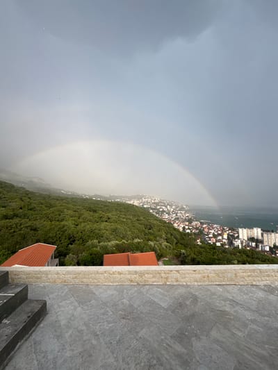 The image depicts a panoramic view from an elevated vantage point, showcasing a coastal city nestled amongst lush green hills and bordering a calm body of water. A faint rainbow arcs across a hazy, overcast sky, suggesting recent or ongoing precipitation. The city is characterized by a dense cluster of buildings with white facades and terracotta roofs, rising up the hillsides and extending towards the waterfront. Several larger, modern buildings are visible along the coast.
In the foreground, a modern stone patio area with steps leads down towards the cityscape. The patio is paved with large, grey stone tiles and a border of smaller, lighter-colored rectangular stones. The overall scene suggests a temperate climate and a scenic, possibly Mediterranean, coastal location. The time of day appears to be late afternoon or early evening, given the soft, diffused light and the overcast conditions. There are no people or specific objects of focus in the image, other than the natural and architectural elements of the landscape. The mood conveyed is one of serene beauty, with the rainbow adding a touch of wonder to the scene. No visible text is present in the image. The location context provided is Sušćepan, Montenegro.