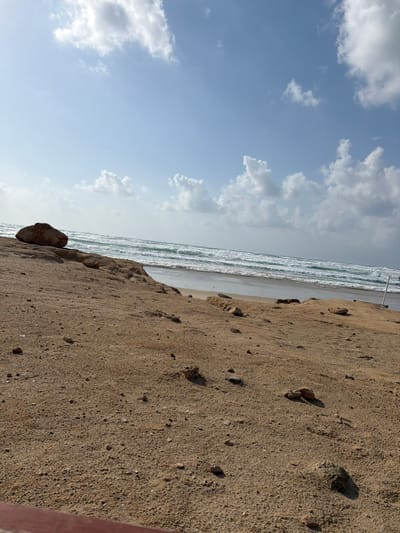 The image depicts a coastal scene at Beit Yanai, Israel, under a partially cloudy blue sky with scattered white cumulus clouds. The sun is visibly bright in the upper left quadrant. The middle ground features the Mediterranean Sea with multiple lines of white-capped waves breaking and receding onto a sandy beach. The foreground and midground are comprised of coarse, light brown to tan sand, exhibiting undulations and containing numerous scattered small, dark-colored rocks and pebbles. A large, dark brown, rounded boulder is positioned on an elevated sandy section in the far left of the frame. On the far right, a slender, light-colored pole extends vertically, partially submerged at the water's edge. A horizontal, reddish-brown object is partially visible at the bottom left corner of the image. No individuals are present, and no specific human actions are discernible within the frame.