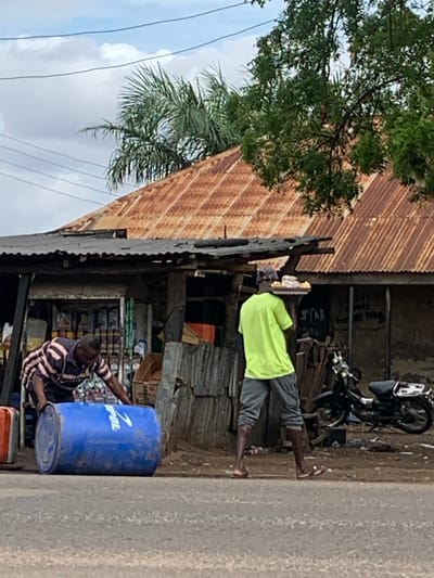 The image depicts an outdoor scene in Shendam, Nigeria, under a partly cloudy sky. In the foreground, a grey paved road stretches across the lower portion of the frame.
On the left side of the road, a male individual with dark skin, wearing a horizontal striped shirt (dark and light beige/pink stripes) and dark trousers, is bent over, pushing a large blue plastic barrel lying on its side. The barrel has white lettering, partially visible as "POL".
Towards the right, a second male individual with dark skin is walking away from the viewer. He wears a bright lime green short-sleeved t-shirt, grey capri-length shorts/trousers, and a dark baseball cap. He is balancing a flat, dark-colored tray on his head, laden with multiple light-colored items, consistent with baked goods or pastries. He wears open-toed footwear.
Behind the two individuals, on an unpaved dirt shoulder, stands a small, open-front structure resembling a shop or kiosk, featuring a dark, corrugated metal roof. Shelves stocked with various products are visible inside this structure. Further back and to the right, a dark-colored motorcycle is parked on the dirt.
The background features several buildings with steeply pitched, rusted corrugated iron roofs. Lush green foliage from various trees, including palm fronds on the left and dense leafy trees on the right, partially obscures these structures. Multiple dark power lines crisscross the light blue and white cloudy sky in the upper left quadrant of the image.