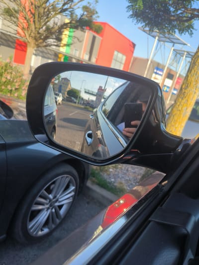 The image shows a reflection in a car's side mirror, capturing a street scene. The mirror itself is black, with a slight crack visible on its surface. Inside the reflection, a person's hand holding a black smartphone is clearly visible, suggesting the photo was taken by the person in the car.

The reflected scene shows a road with other vehicles, including a white car. The background of the reflection features buildings with colorful facades – primarily red, with yellow and green stripes. Trees with green foliage are also present, with one particularly prominent tree in the foreground of the mirrored view. The bright sunlight suggests it is daytime, likely late afternoon given the warm tones.

In the foreground of the main image, the side of a dark-colored car is visible, showing its wheel and part of the door. The setting appears to be an urban or suburban environment, possibly near a commercial area given the building architecture. The overall impression is a candid snapshot taken from inside a vehicle, focusing on the view reflected in the side mirror. There is no visible text in the image or its reflection.