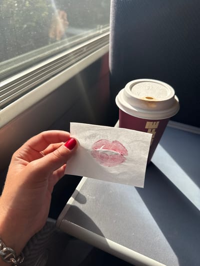 An image taken inside a train or similar vehicle depicts a hand holding a piece of paper with a lipstick print, alongside a coffee cup. The scene is illuminated by sunlight entering through a window on the left.
In the foreground, a human hand, likely female, with red manicured fingernails, holds a small rectangular piece of white paper. The paper bears a distinct impression of red lipstick lips, showing texture and varying pigment density. A silver-colored chain-link bracelet is visible on the wrist. To the right of the paper, a burgundy-colored disposable coffee cup with a white plastic lid rests on a surface. The lid has a small drinking aperture and shows faint brown staining. Partial white lettering, possibly "FEI", is visible on the cup's side.
The background features a window on the left, revealing bright, diffused sunlight and blurred green foliage outside, indicating movement. The window frame is light gray. To the right of the window, a dark blue or gray upholstered seat back is partially visible. Strong sunlight casts defined shadows across the interior surfaces. This scene is located in Assevillers, France.