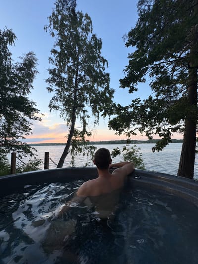 A man is relaxing in a hot tub overlooking a body of water, possibly a lake, at dusk. The sky is a pale blue with streaks of pink and orange from the setting sun. Trees with dark green leaves frame the scene on either side, with one tall birch tree standing prominently in the middle. The water in the hot tub is dark and rippling, reflecting the sky. The man is seen from behind, with his arms resting on the edge of the tub. The location is Priedesmuiža, Latvia.