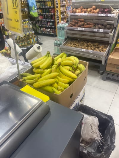 The image appears to be taken inside a grocery store, possibly in Гожули, Ukraine, given the context. In the foreground, a metal scale platform and a grey surface are visible, likely part of a checkout counter. To the right, a large cardboard box is overflowing with ripe bananas. Next to the box is a black trash bag containing white plastic bags. In the background, shelves are stocked with various grocery items, including bottles of oil, bags of produce like potatoes, beets, carrots, and onions, and packaged goods. A price tag indicating "90 кг" (90 kg) is also visible in the upper left corner, suggesting a pricing or weighing station. The lighting is bright, typical of a retail environment. No people are directly visible in the frame.