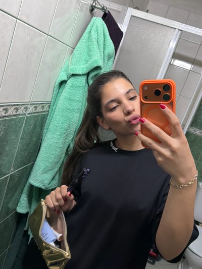 A young woman takes a selfie in a bathroom. She holds an orange smartphone up to her face, pouting at the camera. She wears a black shirt with white lettering at the collar and a gold bracelet. Her fingernails are painted red. In her other hand, she holds a black tube and a gold makeup bag filled with cosmetic products. A green towel hangs next to her on a towel rack. The bathroom has white and green tiled walls and a white shower door is partially visible. The location is Rudartsi, Bulgaria.