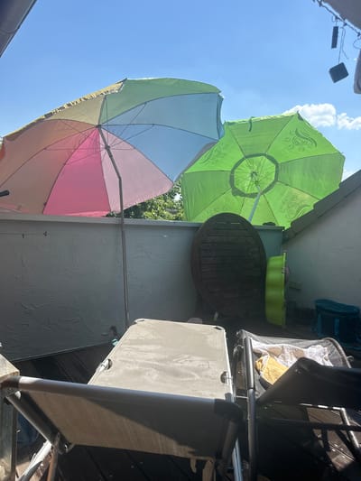 The image depicts an outdoor patio area, likely in a residential setting. The perspective is from below, looking upwards towards a bright blue sky with some scattered clouds, suggesting it is daytime. The scene is dominated by two large patio umbrellas. One umbrella is a vibrant lime green with a patterned design, while the other is a colorful umbrella with sections of pink, light blue, and green.
Several objects are visible on the patio. There is a round, dark wooden table positioned between the two umbrellas. To the right of the table, there is a light green plastic tub. Further to the right, several outdoor chairs are present. One appears to be a lounge chair with a grey mesh fabric and a metal frame, partially folded. Another chair, possibly made of darker material, is also visible. In the background, a wooden deck or flooring is visible, and a grey textured wall forms the backdrop. Some foliage from trees can be seen peeking out from behind the umbrellas and wall. A dark bag is also hanging on the wall in the upper right corner.
The overall impression is of a relaxed outdoor space, possibly prepared for leisure activities or for providing shade on a sunny day. There is no visible text in the image. The lighting suggests it is a sunny afternoon.