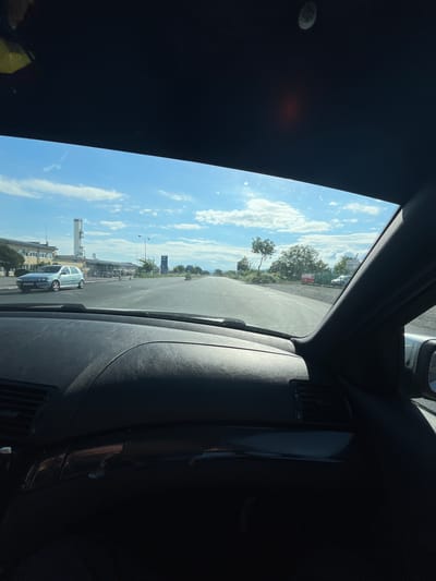 The view from inside a car looking out the windshield onto a street in Kalitinovo, Bulgaria. The sky is bright blue with scattered white clouds. Buildings, trees, and a few cars are visible in the distance. The car is parked on the side of the road. There are no visible people.