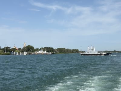 The image shows a wide river with two ferries on it. The ferry on the left is docked at a pier on the left side of the image, and appears to be a car ferry. The ferry on the right is in motion, moving away from the viewer towards the right side of the image. The river is a deep blue-green color, and there are white wakes behind the moving ferry. The background consists of a treeline, buildings, and a clear blue sky with some wispy clouds. The overall scene suggests a sunny day. The location is the Rhone River in Arles, France, where ferries like these are used to transport vehicles and passengers across the river.