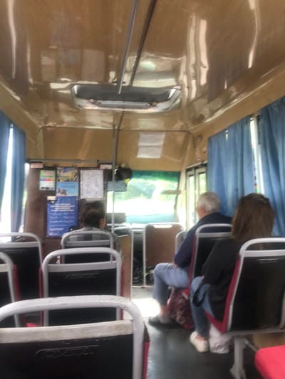The image shows the interior of a bus, likely in Rivnopillia, Ukraine, with several passengers seated. The bus has a tan-colored ceiling and blue curtains covering the windows. In the foreground, several empty seats are visible, some with red trim. Further back, passengers are seated, with their backs to the camera. One passenger in the middle has gray hair and is wearing a blue shirt and gray pants. To their right, a passenger with brown hair is wearing a black top and blue jeans. In the background, there are informational posters mounted on the wall of the bus. The overall impression is of a typical bus interior during a journey.