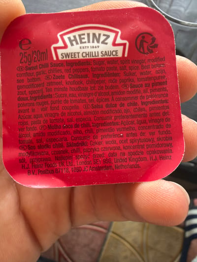 A close-up shot captures a person's hand holding a small, red Heinz Sweet Chilli Sauce sachet. The hand's thumb and fingers are prominently featured, displaying visible skin texture and fingerprints. The sachet is slightly bent as it's held.
The packaging is red with white and black text. The "HEINZ" logo with "ESTP 1869" is visible, along with "SWEET CHILLI SAUCE" and "25g/20ml." A recycling symbol (a person dropping an item into a bin with arrows) is printed near the top left of the sachet.
Extensive product information is printed on the sachet in multiple languages, including English, Dutch, French, Spanish, Portuguese, and Polish. Key visible text includes "Sweet Chilli Sauce. Ingredients: Sugar, water, spirit vinegar, modified cornflour, garlic, chillies, red peppers, tomato paste, salt, spice. Best before: see bottom." Similar ingredient lists are provided in other languages. Manufacturer details are also present: "H.J. Heinz Foods UK Ltd., London SE1 9SG, United Kingdom. H.J. Heinz B.V., Postbus 87118, 1080 JC Amsterdam, Netherlands."
The background is blurred, showing indistinct patterns that suggest an indoor setting, possibly fabric or upholstery. There are no discernible time of day, weather, or emotional cues. The image focuses almost exclusively on the hand and the product packaging.