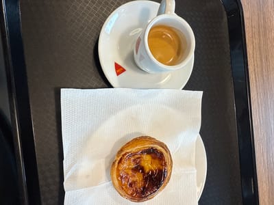 On a black tray, an espresso in a white "Delta" cup and saucer is positioned below a Portuguese custard tart (Pastel de nata) served on a small plate with a white paper napkin. The items appear to be situated on a table with light-colored wood grain visible on the left edge of the image. The location is Pombal, Portugal.