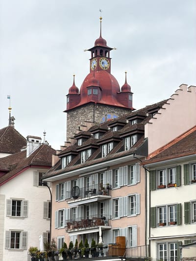 The image shows a section of the city of Lucerne, Switzerland. A distinctive red-roofed clock tower, likely the Zytturm, dominates the left side of the frame. The tower's clock face is visible. Adjacent to the tower are buildings with traditional European architecture, featuring rows of windows with shutters and steeply pitched roofs. Balconies with greenery are visible on the buildings. The sky is overcast.
