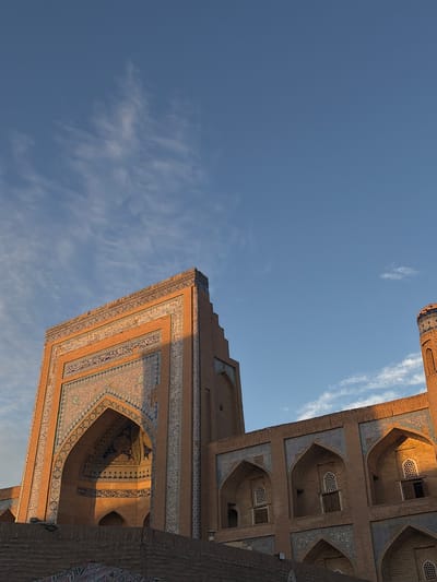 The image shows a close-up, low-angle view of the Kalta Minor Minaret and the adjacent Madrasah in Khiva, Uzbekistan, under a clear blue sky with scattered wispy clouds. The iconic turquoise-tiled minaret is partially visible on the right, catching the warm golden light of what appears to be sunset. The building's facade is constructed from weathered brick, adorned with intricate blue and white geometric and calligraphic tilework, especially around the arched windows and the base of the minaret. The main structure in the foreground features a large, ornate archway, also decorated with detailed tilework, leading into a darker interior space. The architectural style is characteristic of Central Asian Islamic design, with pointed arches and decorative patterns. The lighting suggests either early morning or late afternoon, casting long shadows and highlighting the textures of the brick and tiles. No people are visible in the frame, and the scene is static. The foreground shows the top of a lower brick wall, with some patterned fabric visible underneath.