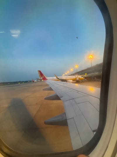 The image is taken from an airplane window, looking out at an airport tarmac during dusk. The sky is a gradient of blue and gray, with the lights of the airport beginning to illuminate the scene. In the foreground, a portion of an airplane wing is visible, with its characteristic metallic panels and rivets.
Further out on the tarmac, several airplanes are parked. The most prominent is a yellow airplane with a cartoon duck logo on its side and the words "Air Travel" and Chinese characters on its fuselage. This plane is connected to a jet bridge. Another airplane with a red and gold tail fin is visible in the middle ground. In the background, airport terminal buildings and rows of illuminated light poles are present, casting a warm glow. The tarmac itself is paved and appears to be marked with lines. The overall impression is that of an airport at a transitional time of day, with planes preparing for or having just completed flights. The location is identified as Xinwu District, China.