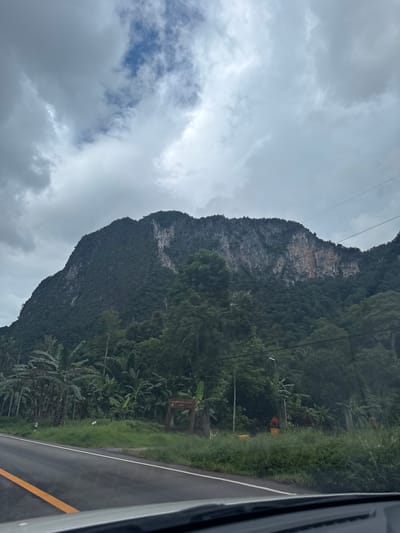 The media file shows a mountainous landscape with lush green vegetation and a cloudy sky. A paved road with yellow and white lines runs along the right side of the frame. The mountain is steep and covered in trees, with a bare rock face visible on the left side. There are also some power lines visible against the sky. In the background, there are several banana trees and other tropical foliage. There are a few small signs visible, one of which appears to be a wooden signpost. The overall impression is of a natural and somewhat remote location, possibly a roadside view in a tropical region. The weather appears overcast, suggesting it might be a cloudy day or approaching rain. The time of day is difficult to determine due to the overcast conditions, but it appears to be daytime. There are no people visible in the image. The scene evokes a sense of natural beauty and tranquility, with the road suggesting human presence and access to this area. The location context of Ban Tam Tong Lang, Thailand, further reinforces the tropical and mountainous setting.