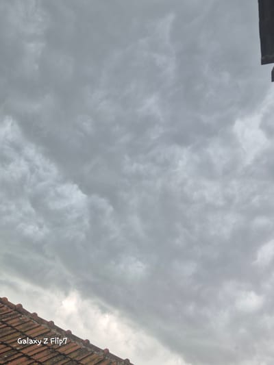 The image shows a cloudy sky with a dark object at the top left and a section of a terracotta tiled roof at the bottom right. The photo was likely taken in Kalipetrovo, Bulgaria. The sky is overcast and gray, suggesting a rainy or gloomy weather. The roof tiles appear aged and may be part of a building. The “Galaxy Z Flip7” watermark is on the bottom right of the image.