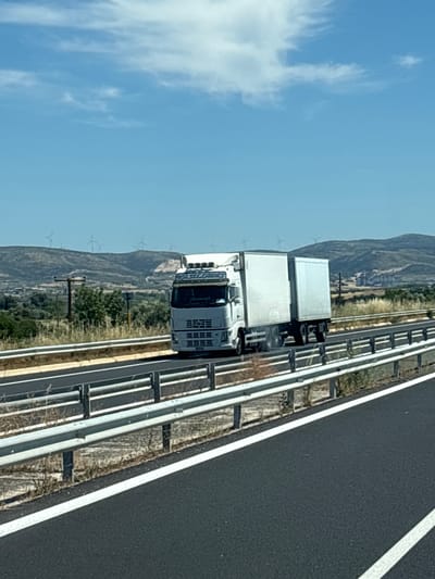 A white semi-trailer truck with a two-part white trailer is driving on a multi-lane highway in a rural area under a clear blue sky with scattered clouds. The truck is positioned in the right lane, moving away from the viewer and slightly to the left. The truck's cab has several lights on the roof and a visible grille. The trailer appears to be a standard refrigerated unit.
The highway is bordered by a metal guardrail on the right side, with dry vegetation and some scattered scrub brush growing on the shoulder. The road surface is asphalt, with white lane markings visible. In the background, a hilly landscape is visible, with several wind turbines scattered across the hilltops. Some trees and low vegetation are also present on the slopes. Further in the distance, there appear to be some buildings or industrial structures on a hillside. The scene suggests a sunny day with good visibility. The specific location within Alexandroupolis, Greece, is not discernible from the visual elements of the image alone.