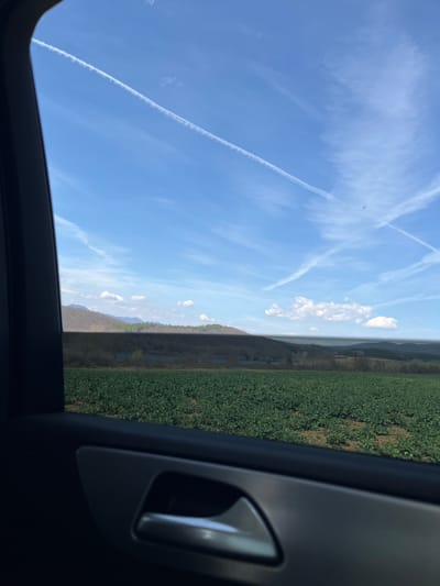 The image depicts a rural landscape near Ruska Bela, Bulgaria, viewed from the interior of a vehicle through a side window. The left portion of the frame is occupied by the dark gray door frame and a metallic silver door handle. Beyond the window, a vast field of dense, dark green low-lying crops covers the immediate foreground and midground. Visible within this field are intermittent patches of light brown soil. A dark, shadowed band of vegetation, possibly obscuring a body of water, stretches horizontally behind the field. In the background, rolling hills and distant mountains, some with visible tree cover, rise towards the horizon. The sky is bright blue with scattered white cumulus clouds and multiple distinct white contrails from aircraft, some crossing. No individuals, direct actions, or interactions are visible.