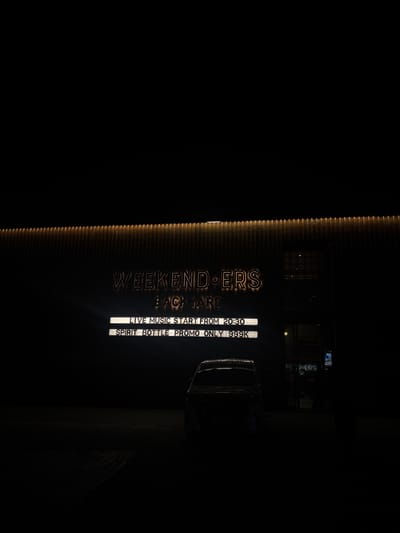The image depicts the exterior of a building at night in Babakan Madang, Indonesia. A neon sign on the building reads "WEEKENDERS BACKYARD" in illuminated lettering. Below this, two rows of illuminated text display event information. The top row states "LIVE MUSIC START FROM 20:30", and the bottom row reads "SPIRIT BOTTLE PROMO ONLY 999K". A dark-colored car is parked in front of the building, partially silhouetted against the lit signage. The surrounding environment is dark, suggesting it is nighttime. The building's facade features vertical ribbed detailing illuminated from above.