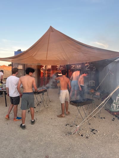 The image shows a group of five men gathered beneath a large canopy in Tze'elim, Israel. The men are standing around several charcoal grills, cooking and smoking. The setting is a desert landscape. Two of the men are wearing shorts and flip-flops, while the others are shirtless and wearing shorts or swim trunks. One of the men is holding a grill spatula. The canopy is made of brown canvas and is supported by metal poles. In the background, a metal container labeled with the word “ZIM” is partially visible. The ground is composed of gravel and sand. The sky is clear and blue. The time appears to be early evening, as the light is warm and the shadows are long. The scene evokes a sense of relaxation and camaraderie. The men are enjoying the outdoors and the process of cooking together.