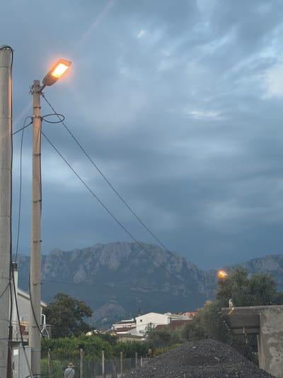 This image, taken in Burtaiši, Montenegro, depicts a street scene under a cloudy sky. Two concrete utility poles dominate the foreground, with the pole on the left supporting a visible bundle of wires and electrical conduit. The pole on the right is taller and supports a single streetlamp that is illuminated with a warm, orange-yellow light. Overhead, power lines crisscross the frame, strung between the poles. In the background, a range of jagged mountains forms a dark, imposing silhouette against the muted sky. Below the mountains, a scattering of houses and trees are visible, suggesting a residential area. A pile of dark gravel is situated in the lower right portion of the image, and a portion of a rough concrete structure is also visible. A person wearing a hat is partially visible in the lower left corner, walking along a path.
