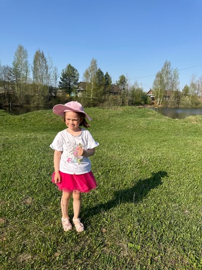 A young girl wearing a pink hat, a white shirt with a unicorn design, and a pink tutu is standing on a grassy lawn. The sky is blue and clear. In the background, trees and a body of water are visible. The location is near Черёмушки, Russia.