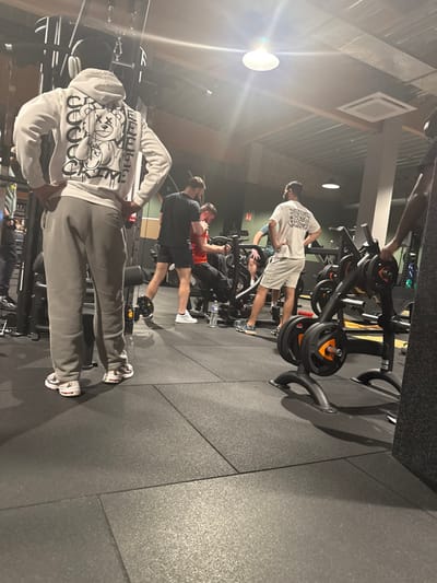 The image captures an indoor scene within a gym or fitness center, characterized by dark grey, textured rubber flooring and various exercise equipment. The lighting is artificial, with bright overhead lights creating some glare and highlights in the image. The perspective is from behind and slightly to the left of the main subject.
In the immediate foreground, a man is visible from the waist down, and then from the back upwards. He is wearing a light grey hooded sweatshirt adorned with a prominent black and white graphic design on the back, featuring several cartoon-like characters (resembling bears or stylized figures with X'd out eyes) and some partially visible stylized text, possibly including "GOOD" or "GOOCH" and other symbols. He is also wearing light grey sweatpants and white sneakers with red and blue accents. His hands are placed on his hips, and he appears to be wearing white over-ear headphones.
In the midground, several other men are engaged in activities or resting. To the right of the foreground man, another man is standing next to a gym machine. He is wearing a black t-shirt, black shorts, and white sneakers. A white water bottle is placed on the floor near his feet. Further back and slightly to the right, a third man is seen using a gym machine, possibly a rowing machine or a chest press, with his back mostly turned to the camera. He is wearing a red t-shirt.
In the background, another man is standing upright near a weight rack or machine. He is dressed in a white t-shirt with an unidentifiable graphic design on the back, light-colored shorts, and white flip-flops or slides. His focus appears to be on the equipment in front of him. Various black gym machines with attached weight plates (black with orange accents) are scattered throughout the space. The ceiling features exposed beams or panels, and a darker wall is visible further back. The overall atmosphere suggests a typical gym environment with individuals exercising or preparing to exercise.