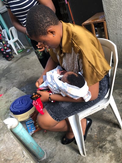A woman sits outdoors on a white plastic chair, holding a baby wrapped in a patterned cloth. She is dressed in a gold top and dark shorts. The baby is positioned against her chest. Next to the woman is a blue and cream-colored object, and further away is a blue pipe. The ground is concrete. Additional chairs and objects are scattered around the area.