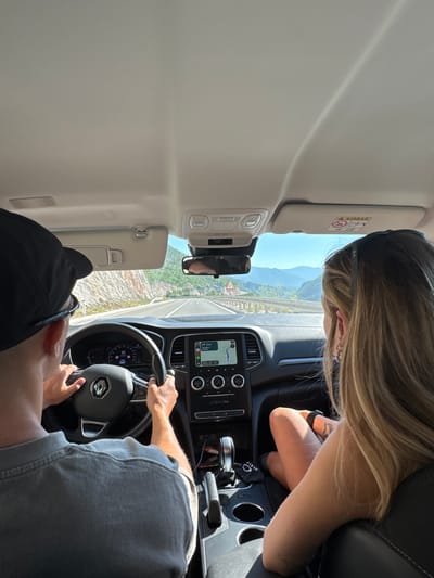 The image shows two people inside a car, seen from the back seats. The person on the left is driving and wearing a black baseball cap and a grey t-shirt. The person on the right is a woman with blonde hair, wearing a black sleeveless top and jewelry on her wrist and ears. She is looking out the window at a mountainous landscape. The car's dashboard is visible, including a navigation screen displaying a map and a digital speedometer showing 95. The car is on a road in a mountainous area, with a steep rocky incline on the left side of the road and a valley with layered mountains in the distance. The sky is clear and blue. The location is Šipačno, Montenegro.