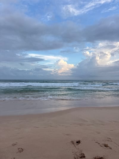 This is a photograph of a beach in Koggala, Sri Lanka. The scene depicts the ocean meeting the shore under a cloudy sky. Gentle waves are breaking on the sandy beach, leaving a wet sheen on the sand. Footprints are visible in the foreground of the sand. The sky is filled with a mix of dark and light clouds, with a patch of blue sky visible in the distance where the clouds are less dense. The overall mood is calm and serene.