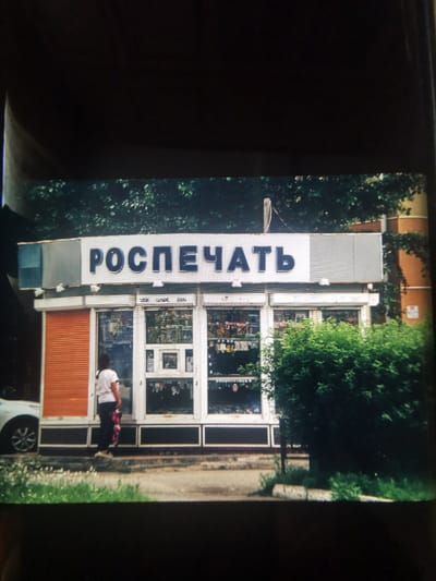 The image depicts a street scene in Serov, Russia. In the foreground, a woman in a white t-shirt and dark pants stands in front of a small kiosk. She is holding a colorful bag. To the left of the kiosk, the front of a white car is partially visible. The kiosk itself has a light-colored facade with a prominent sign above it. The sign displays blue Cyrillic text, which reads "РОСПЕЧАТЬ". Behind the kiosk, lush green trees and foliage create a natural backdrop. The kiosk's front features large glass windows, behind which various items are displayed. One side of the kiosk has orange shutters. The ground in front of the kiosk is paved, with some greenery and wildflowers growing along the edge. The overall impression is of a typical urban street scene, likely during the daytime, given the illumination. The weather appears to be fair, with no obvious signs of rain or harsh sun. The image is taken from a slightly elevated perspective, looking down towards the street.