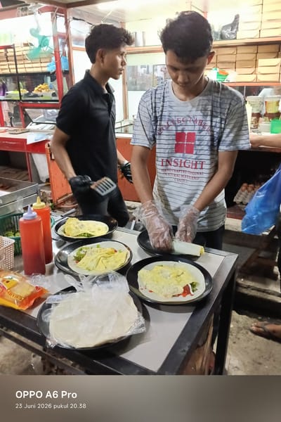 Two young men are preparing food at an outdoor food stall in Sangso, Indonesia. One man, wearing a black polo shirt and black gloves, is holding a spatula and appears to be cooking or serving food on a plate. The other man, wearing a grey and white striped t-shirt and clear plastic gloves, is in the process of wrapping food. In the foreground, there are several plates of food, including what looks like crepes or wraps filled with eggs and vegetables, along with two squeeze bottles of condiments. A stack of what appear to be tortillas or flatbreads wrapped in plastic is also visible. Behind the men, shelves are stocked with boxes and containers, suggesting a busy food preparation area. The lighting suggests it is either dusk or evening. The timestamp at the bottom of the image reads "OPPO A6 Pro 23 Juni 2026 pukul 20.38," indicating the date and time the photo was taken.