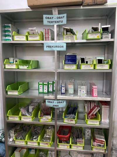 The image shows a well-organized pharmaceutical cabinet filled with various medications. The cabinet is divided into multiple shelves, each containing brightly colored plastic bins and boxes holding blister packs, vials, and other drug packaging. Some bins are labeled with handwritten notes and printed labels, indicating drug names, dosages, and categories such as "OBAT - OBAT TERPECU" (Medicine - Specific Medicine), "PREKURSOR" (Precursor), and "OBAT - OBAT JIWA" (Medicine - Psychiatric Medicine).
Visible medication names include "TRAMADOL," "LAPISIV TABLET," "PARATUSIN TAB," "CLOZAPIN," "HALOPERIDOL," "OLANZAPINE," "FLUOXETINE," and "TRIHEXIPHENIDIL." The setting appears to be a pharmacy or a medical storage area, likely in Margoyoso, Indonesia, given the context provided. The lighting is bright and even, suggesting an indoor environment, possibly during the daytime. There are no people visible in the image. The overall impression is one of order and efficiency in a medical supply setting.