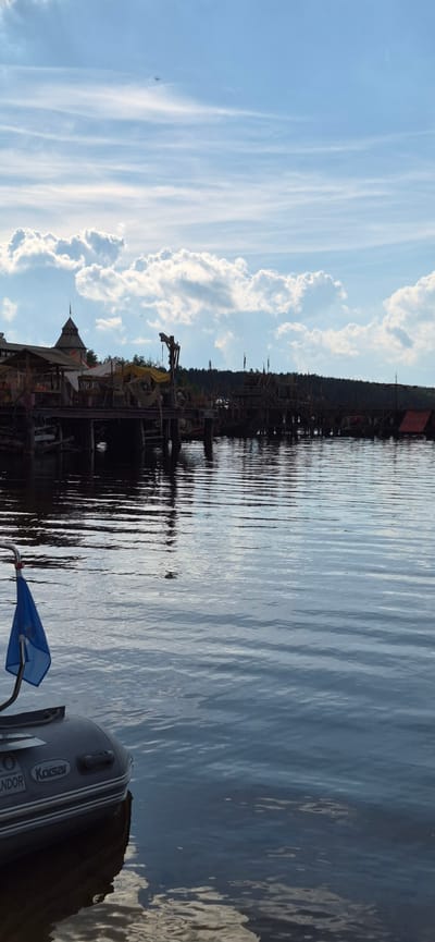 The image shows a body of water with a wooden pier and structures along its edge, likely part of a themed attraction or historical reenactment site, possibly in the city of Моторное, Russia. A small inflatable boat with a blue flag is in the foreground, positioned on the left side of the frame, partially submerged in the water. The boat has branding visible, including "Korsar." The water exhibits ripples, reflecting the sky and the structures on the pier. The sky is partly cloudy with a blue hue. The pier is lined with wooden buildings and what appear to be older boats or ship parts, with some canvas coverings and cranes suggesting a historical or maritime theme. Trees are visible in the background behind the pier. The overall scene depicts an outdoor waterfront environment under a bright sky.