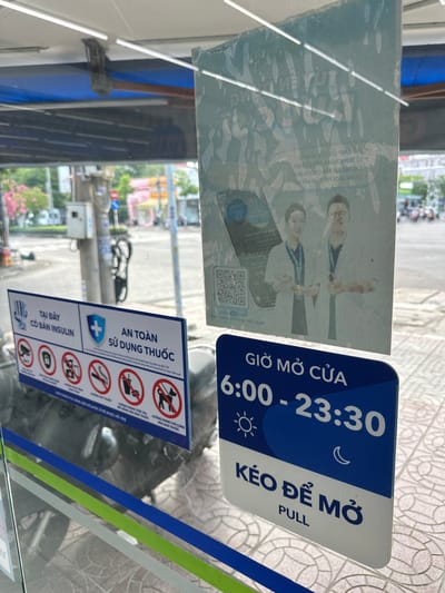 The image shows a close-up of a glass door with various signs and posters affixed to it. In the foreground, a blue and white sign displays business hours, reading "GIỜ MỞ CỬA 6:00 - 23:30" and "KÉO ĐỂ MỞ PULL". To the left of this sign is a rectangular blue and white sign with several icons and text. The top of this sign reads "TẠI ĐÂY CÓ BÁN INSULIN". Below this, there are icons representing a security camera, a person being photographed, a mobile phone with a prohibition symbol, a cigarette with a prohibition symbol, a disposable coffee cup with a prohibition symbol, and a dog with a prohibition symbol. Text below these icons includes "AN TOÀN SỬ DỤNG THUỐC", and other Vietnamese phrases. Reflected in the glass is a street scene with buildings, traffic, and trees. Behind the glass, a motorcycle is partially visible. A larger, faded poster is adhered to the glass above the signs, featuring two individuals in medical attire and some text in Vietnamese.