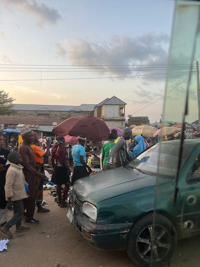 The image depicts a bustling open-air market scene in Rinjim Gani, Nigeria, at approximately late afternoon or early evening, indicated by the warm, transitioning light blue to orange-yellow sky with scattered grey clouds.
In the foreground, a dark green vehicle, consistent with a Volkswagen Golf Mk3, is partially visible on the right, angled towards the left. The car exhibits damage, specifically a missing lens from its front driver's side headlight and damage to the front bumper. Reflections of the sky and surrounding environment are visible on the car's windshield and side window, suggesting the photograph was taken from within another vehicle or behind glass.
The unpaved ground, appearing as dirt or worn earth, is densely populated with people and market activities. Numerous individuals, including adults and at least one child, are present. On the far left, a young boy in a light-colored jacket and dark pants walks. A man in a brown traditional shirt and patterned cap stands in the left-midground, facing right. People wear a variety of attire, including head coverings and casual clothing. A cluster of assorted footwear is laid out on the ground in the mid-ground, indicating a shoe vendor.
Behind the crowd, several market stalls are delineated by large umbrellas providing shade. A prominent dark maroon umbrella is situated in the mid-ground center-left. Other yellow umbrellas bear discernible branding such as "GLO Cafe & POS," "CORRECT VOICE," and "3X POS & DATA." Merchandise is visible under these shelters.
In the background, a series of low-rise buildings with corrugated metal roofs extends across the horizon. One light-colored, more ornate building with a distinctive roof structure is visible on the left side of the mid-background. Overhead, multiple power lines traverse the upper portion of the frame. The overall scene is characterized by active commerce and pedestrian movement.
