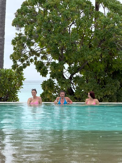 The photograph shows three women in a swimming pool, seemingly an infinity pool, given the ocean backdrop, possibly in Guraidhoo, Maldives. The woman at the top of the image has auburn hair and wears a pink swimsuit. The woman in the middle wears a blue swimsuit. The woman at the bottom has black hair pulled back and wears a pink swimsuit and sunglasses and holds a beverage and straw. The pool edge is adjacent to lush tropical foliage. The water is aqua to green. The sky is overcast.