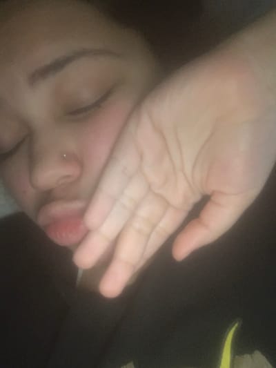 The image displays a close-up, portrait-oriented view of a person, likely female, with eyes closed, suggesting rest or sleep. The individual's left hand is positioned across the lower right side of their face, covering the cheek and jawline, with fingers slightly splayed and the palm visible towards the viewer. A small, light-colored stud piercing is visible on the left nostril. The lips are full and appear slightly pursed. The person is wearing a dark-colored garment, possibly a shirt or sweatshirt, with a faint yellow detail visible in the lower right corner of the frame. The lighting is dim and soft, indicating an indoor environment with an indistinct, dark background. The image provides no identifiable features or landmarks to determine the location in the city of Lancaster, United States of America.