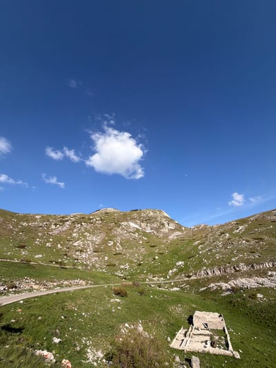 The image shows a landscape of a mountain in Boričje, Montenegro. The view is composed of a blue sky with scattered clouds occupying the left half and the right half featuring the mountain terrain. A stone structure, possibly ruins, sits on the mountainside amidst the green vegetation. A road cuts through the greenery along the base of the mountain.