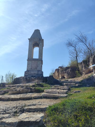A stone monument stands atop a rocky mound in Stroiesti, Moldova, under a blue sky with wispy clouds. The monument is a tall, rectangular structure with an arched opening near its peak, exhibiting weathered stone construction. The mound is composed of rough, light-colored stone steps leading up to the monument, with patches of green grass and small bushes scattered around. To the right of the monument, a rocky cliff with bare trees and some greenery is visible.