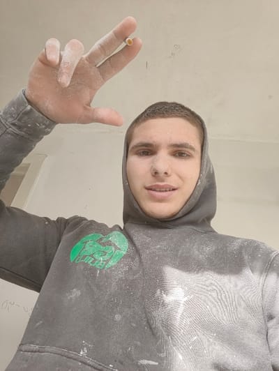 An upward-angled photograph depicts a young adult male, presumably in Deerlijk, Belgium, within an interior room under construction or renovation. The male has short brown hair and is wearing a dark grey hooded sweatshirt with the hood pulled up, featuring a green logo on the left chest. His face, hair, and clothing are significantly coated with a fine white powdery substance, consistent with plaster, drywall dust, or similar construction materials. He looks directly at the camera with a neutral to slight smiling expression. His right hand is raised to the upper left of the frame, also covered in the white powder, holding an unlit cigarette between his index and middle fingers. The background consists of plain, light-colored walls and a ceiling, which also appear unfinished or recently worked on. Faint handwritten markings, such as "320" and "GPL," are visible on the ceiling and a wall.