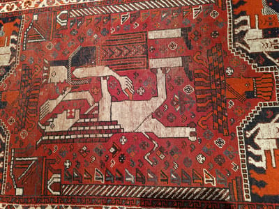 This is a close-up, overhead shot of a portion of an antique rug with a predominantly red background. The rug features a central motif of a stylized human figure rendered in white and beige, lying down with its arms outstretched. The figure is depicted with a white upper body and legs, and a black headdress. The surrounding design is intricate and geometric, with patterns in black, white, orange, and various shades of red and brown. There are smaller, repeating motifs scattered across the rug, including what appear to be floral or star-like shapes. The rug's texture is visibly coarse, indicating it is likely woven from wool. The image does not provide any information about the time of day, weather, or specific location beyond its general association with Hovden, Norway. No text is visible in the image. The overall impression is one of an aged, decorative textile with a rich history.
