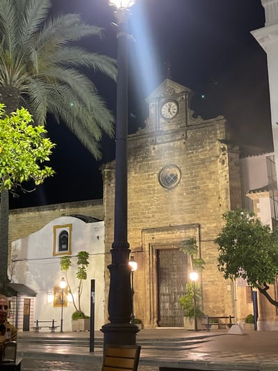 The image shows a nighttime view of a section of Jerez De La Frontera, Spain, featuring a building with a clock tower. A lamppost stands prominently in the foreground, partially obscuring the view. The building is illuminated, with light reflecting off its stone facade. A palm tree and other greenery are visible near the base of the lamppost. In the background, architectural details such as windows, doors, and steps are discernible. A person is partially visible at the bottom of the frame near a table, suggesting an outdoor seating area.