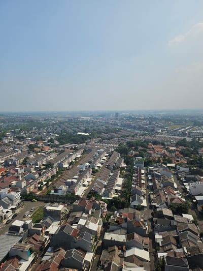 This is an aerial photograph of a suburban residential area in Medan, Indonesia. The scene is dominated by rows of houses with red tile roofs and light-colored walls. The houses are densely packed together, with narrow streets winding between them. There are numerous trees interspersed throughout the neighborhood, adding a touch of green to the otherwise muted color palette. In the background, a hazy skyline of taller buildings suggests the presence of a more urbanized area further out. The sky is a pale blue with a few wispy clouds. The image appears to have been taken on a clear, sunny day. There are no people or visible activity in the photograph.
