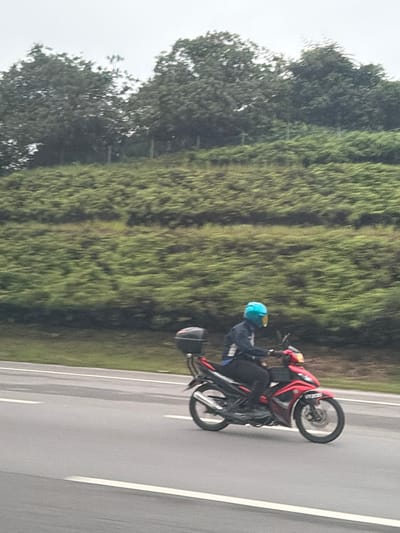 A person wearing a blue helmet and a dark jacket is riding a red and black motorcycle on a wet asphalt road. The motorcycle has a black storage box mounted on the rear. The road has white lane markings. In the background, there is a green, vegetated slope with trees on top. The sky is overcast. The image was taken from a moving vehicle, as indicated by the motion blur. The location is the city of Subang Jaya City Council, Malaysia.